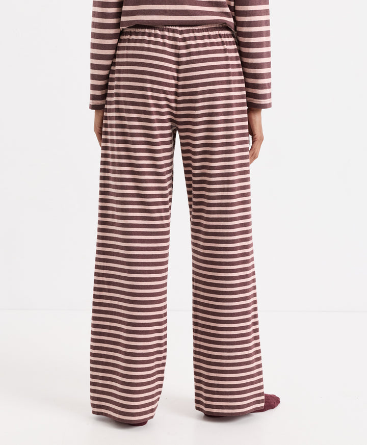 A person shown from the back wearing wide-leg pajama pants with horizontal merlot and clay stripes, paired with a matching striped long-sleeve top, against a plain white studio background.