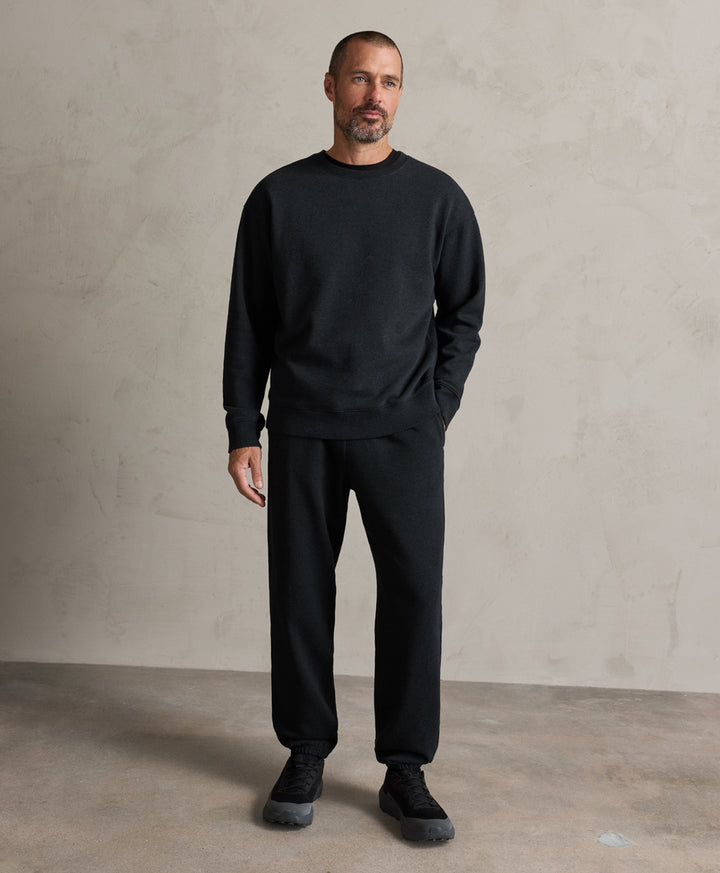 A man wearing a pact charcoal heather crewneck sweatshirt and matching black sweatpants, standing against a textured light gray studio wall.