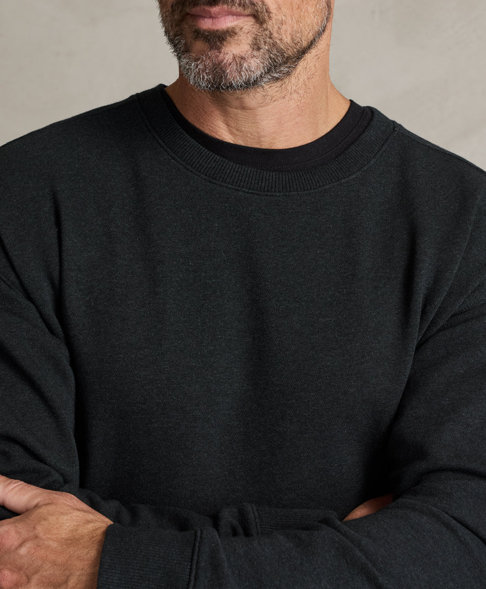 A close-up of a man wearing a charcoal heather crewneck sweatshirt, showing the texture of the fabric and ribbed collar, with his arms crossed.