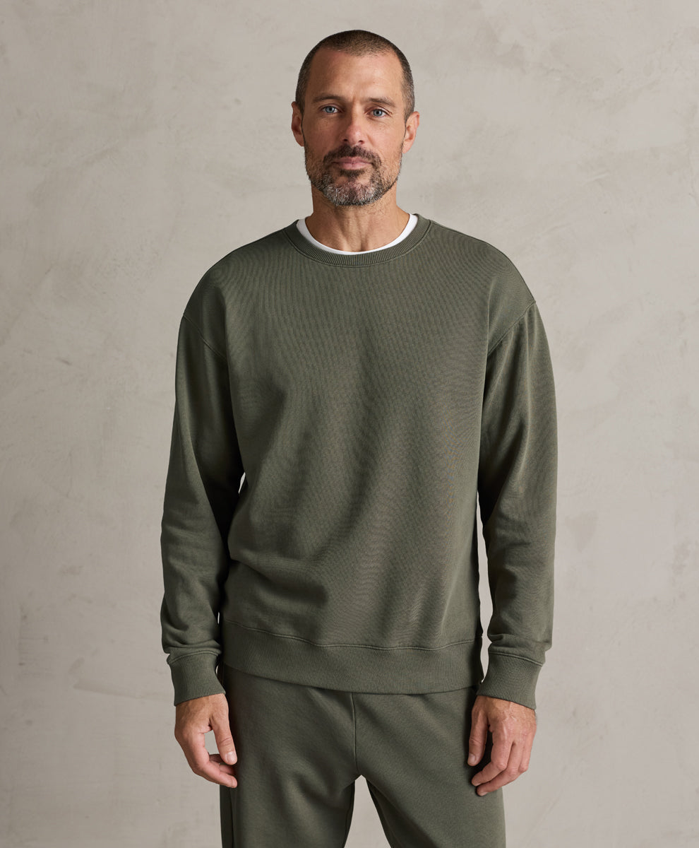 A man wearing a Pact dusted green ribbed crewneck sweatshirt and matching sweatpants, standing against a textured light gray wall.
