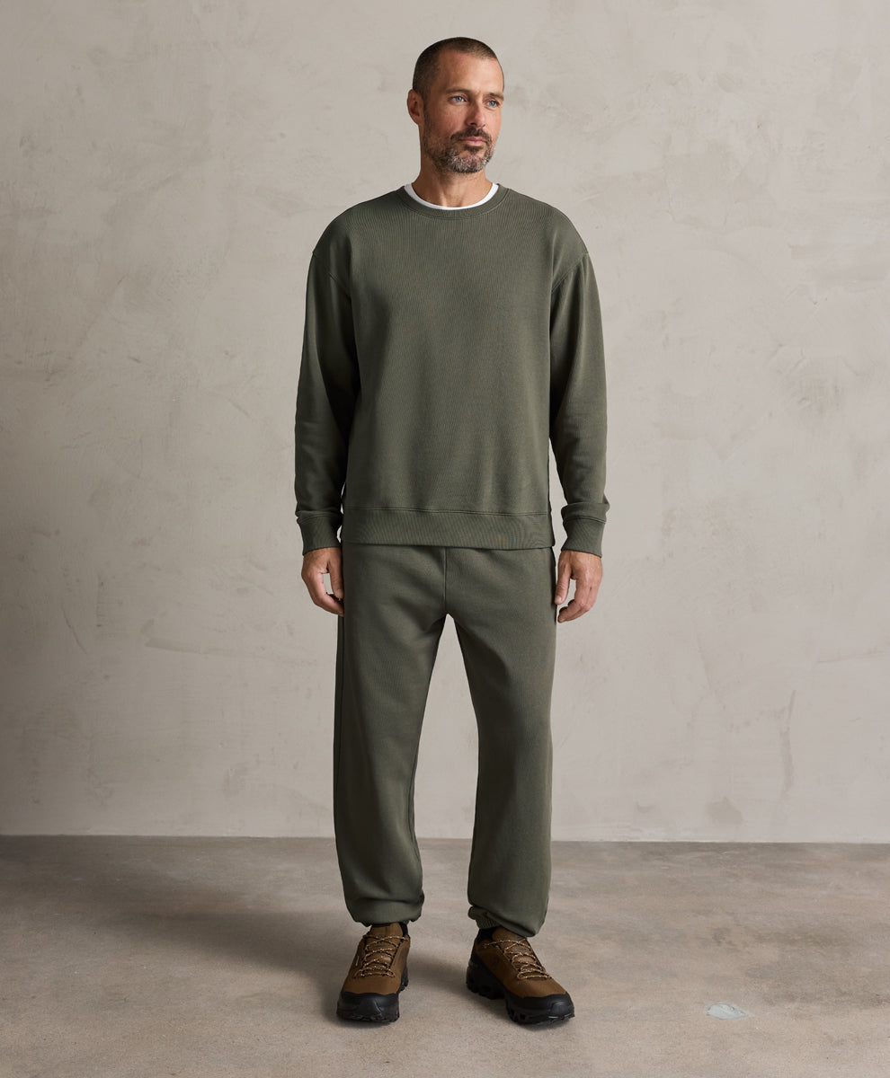 A man wearing a dusted green crewneck sweatshirt and matching sweatpants, paired with brown leather sneakers, standing against a textured light gray studio wall.