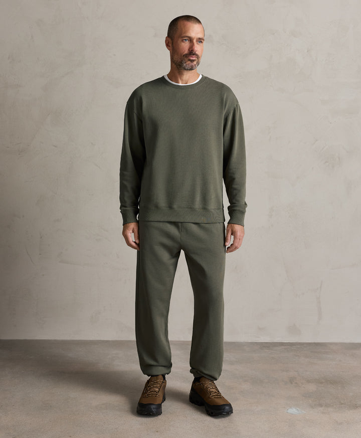 A man wearing a dusted green crewneck sweatshirt and matching sweatpants, paired with brown leather sneakers, standing against a textured light gray studio wall.