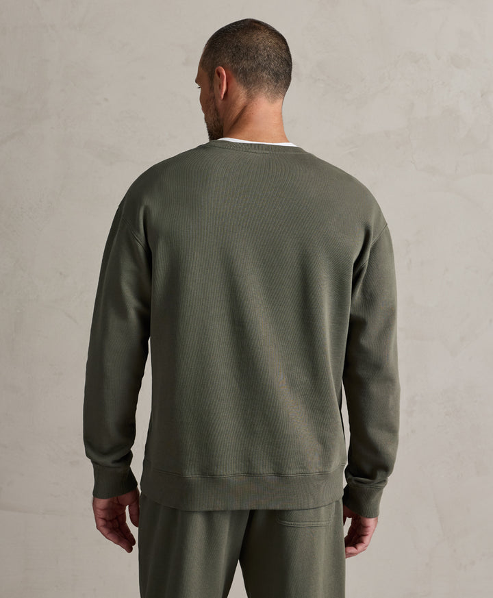 A man viewed from behind wearing an dusted green crewneck sweatshirt and matching sweatpants against a textured light gray wall.