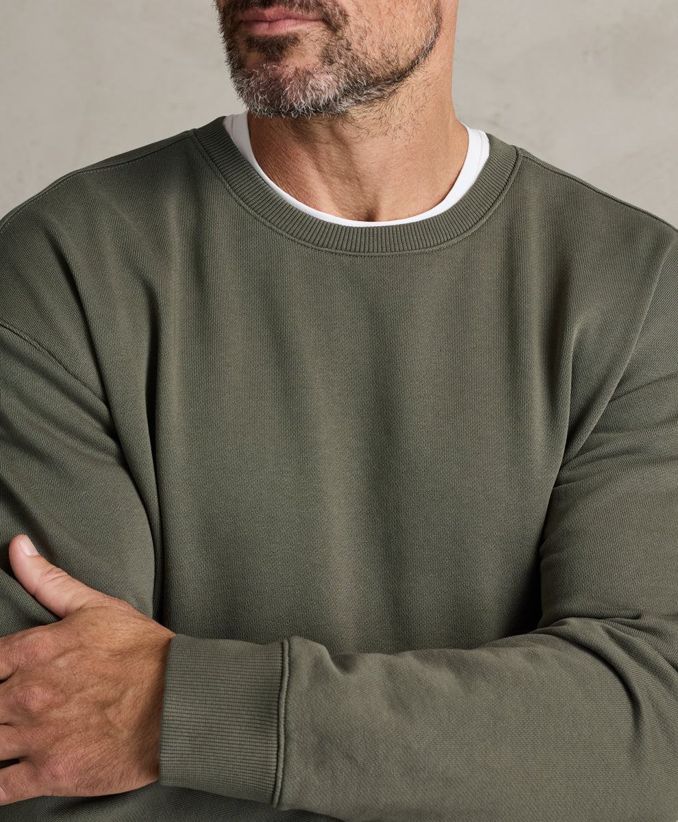 A close-up of a man wearing a dusted green crewneck sweatshirt over a white t-shirt, with his arms crossed against a neutral gray background.