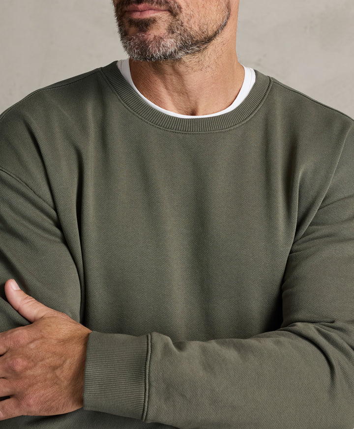 A close-up of a man wearing a dusted green crewneck sweatshirt over a white t-shirt, with his arms crossed against a neutral gray background.
