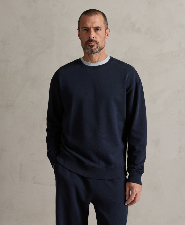 A man wearing a dark navy blue textured knit crewneck sweater and matching navy pants, standing against a textured light gray wall.