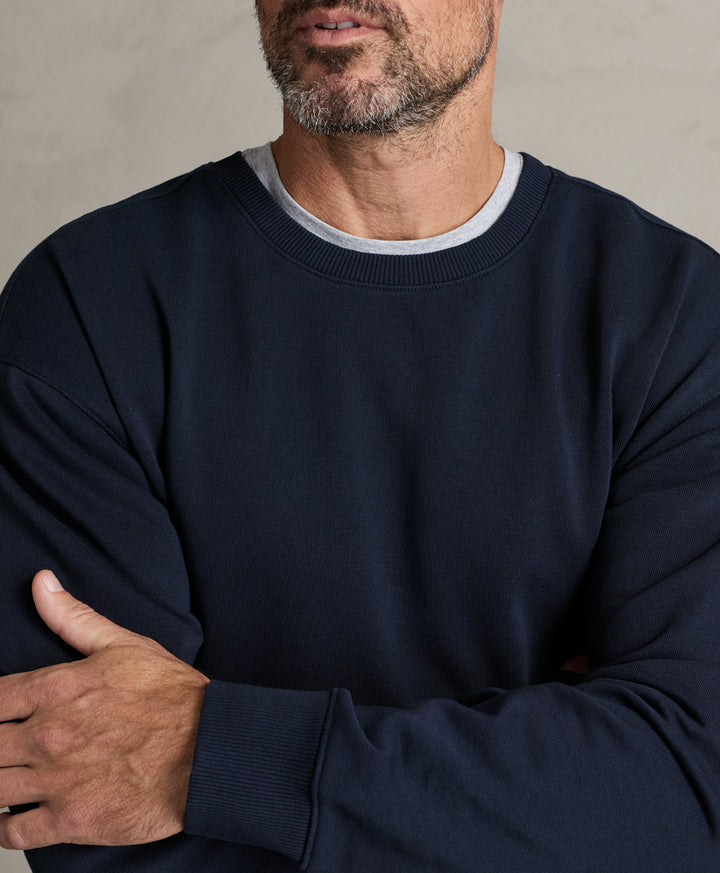 A close-up of a man wearing a dark navy crewneck sweatshirt over a light gray t-shirt, with his arms crossed against a neutral textured background.