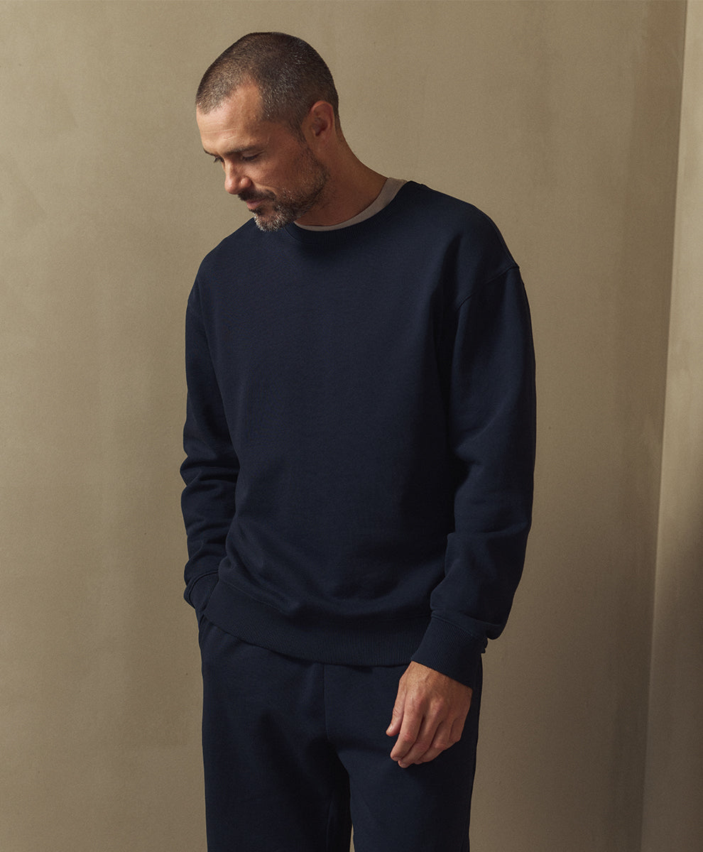 A man wearing a dark navy crewneck sweatshirt and matching navy pants, standing against a textured beige wall.
