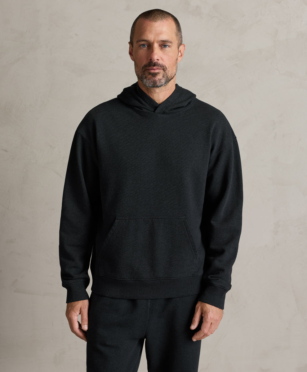 A man wearing a Pact charcoal heather pullover hoodie with a front kangaroo pocket, standing against a textured light gray wall.