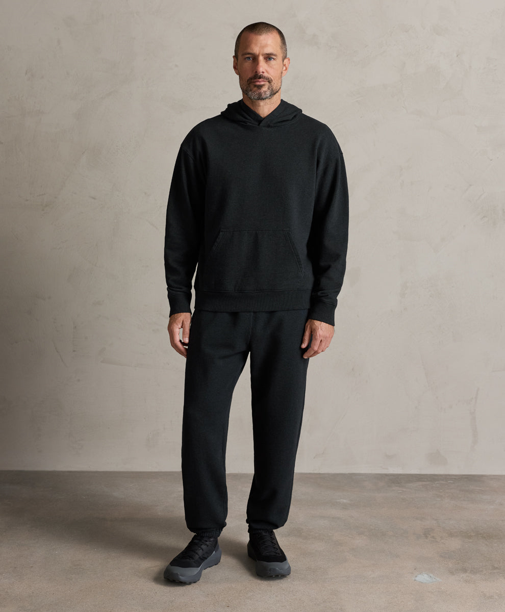 A man wearing a charcoal heather pullover hoodie and matching black sweatpants, standing against a textured light gray concrete wall and floor.