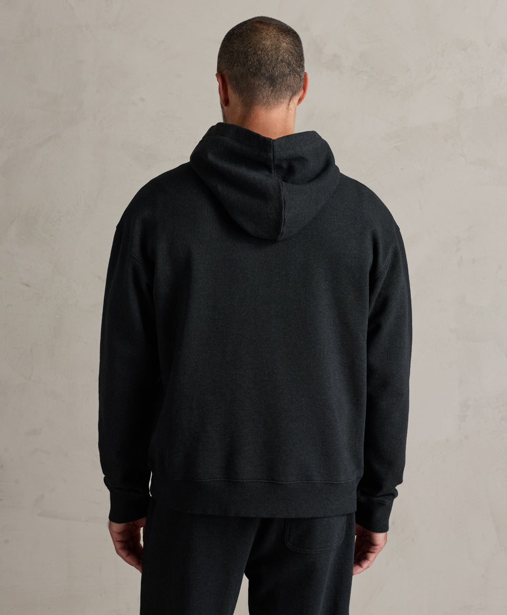 A man viewed from behind wearing a charcoal heather pullover hoodie and black pants, standing against a textured light gray wall.