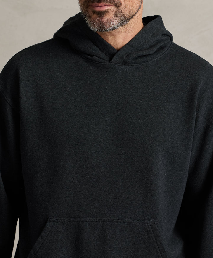 A close-up of a man wearing a charcoal heather pullover hoodie with a front kangaroo pocket, set against a neutral textured background.