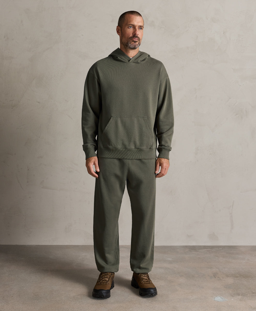 A man modeling a dusted green hoodie and matching sweatpants set, paired with brown lace-up shoes, against a textured light gray studio wall and concrete floor.