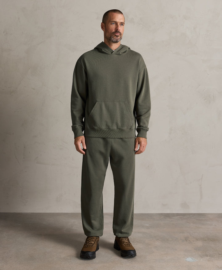 A man modeling a dusted green hoodie and matching sweatpants set, paired with brown lace-up shoes, against a textured light gray studio wall and concrete floor.