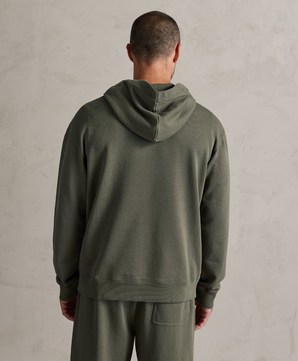 A man viewed from behind wearing a dusted green hooded sweatshirt and matching sweatpants against a textured light gray wall.