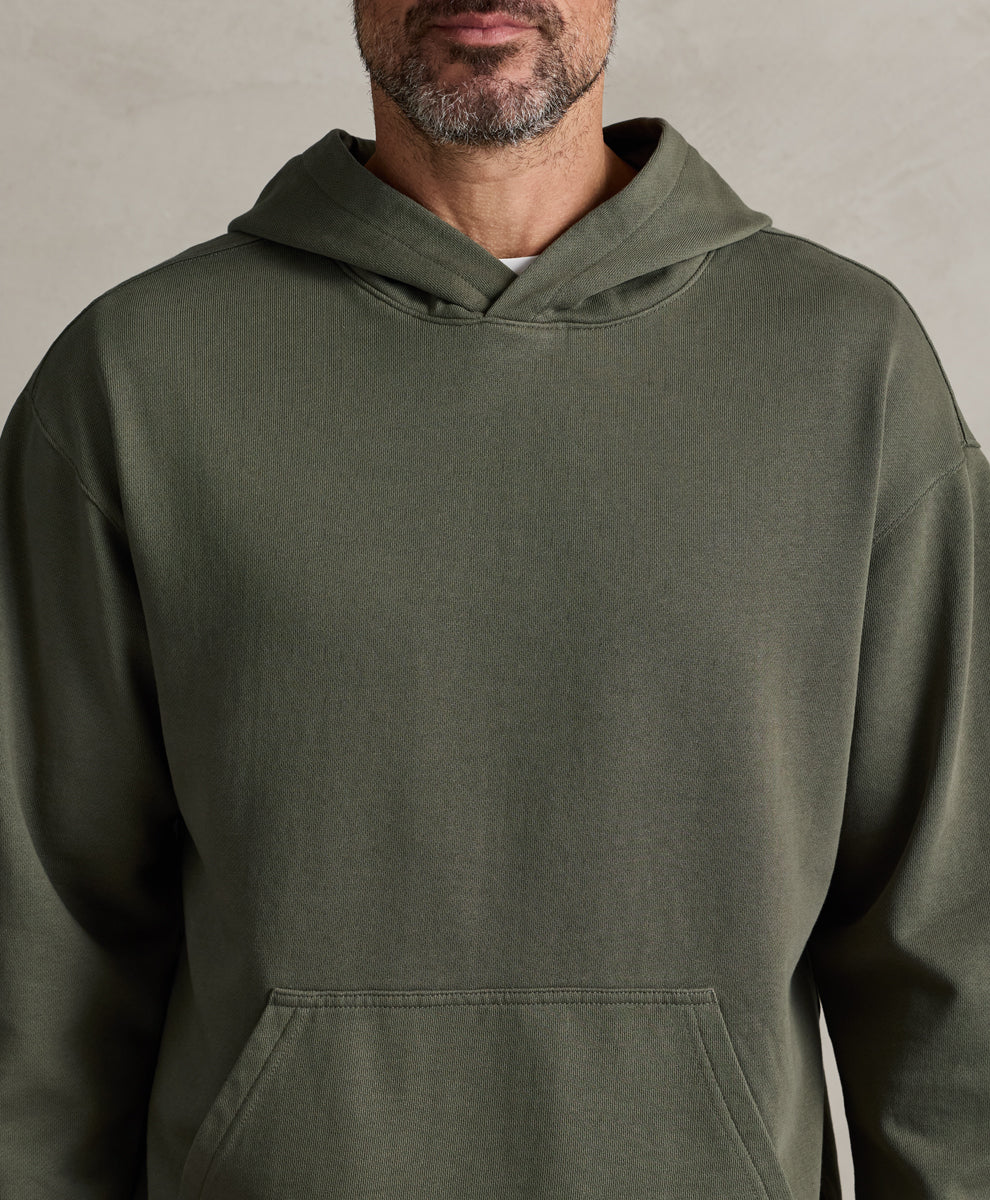 A close-up of a man wearing a dusted green pullover hoodie against a neutral textured gray background.