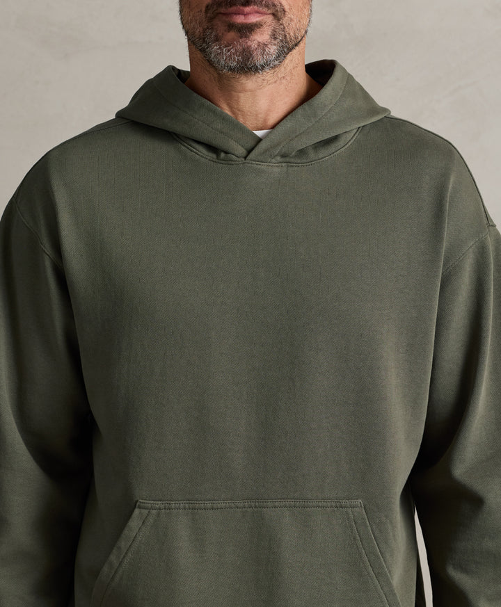 A close-up of a man wearing a dusted green pullover hoodie against a neutral textured gray background.