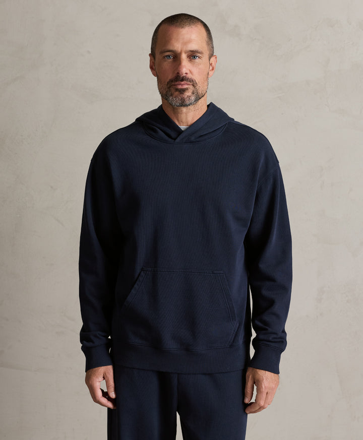 A man wearing a dark navy blue ribbed hoodie sweatshirt and matching sweatpants, standing against a textured light gray concrete wall.