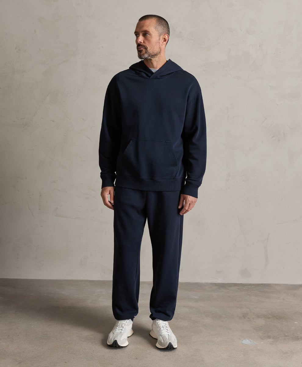 A man wearing a matching navy blue hoodie and sweatpants set, paired with white sneakers, standing against a textured light gray wall.