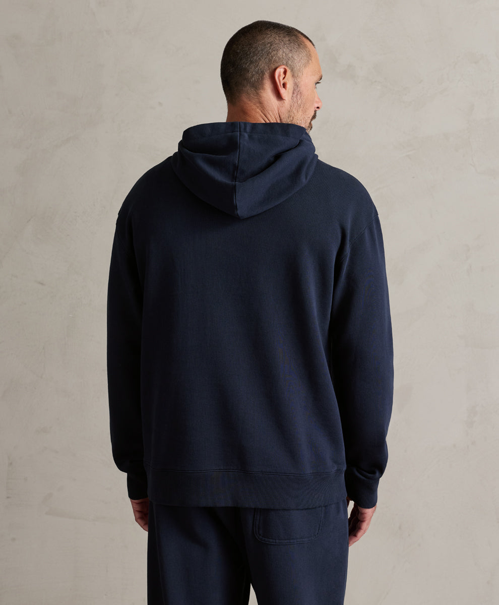 A man viewed from the back wearing a dark navy blue hooded sweatshirt and matching navy pants, standing against a textured light gray wall.