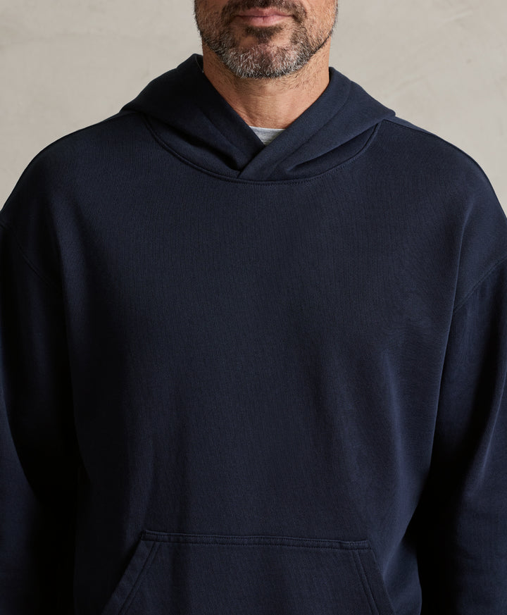 A close-up of a man wearing a dark navy blue pullover hoodie with a kangaroo pocket, shown from the chest up against a neutral textured wall.