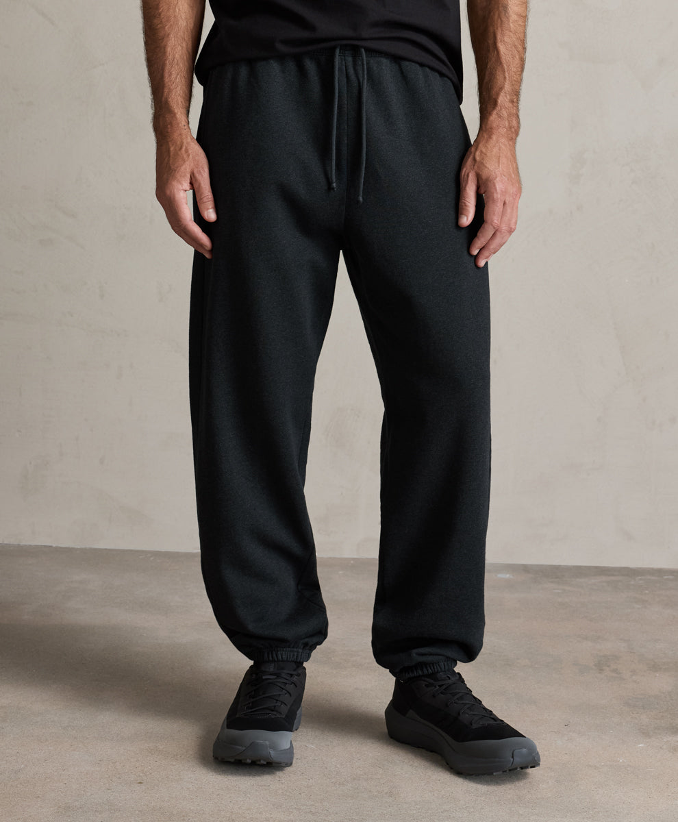 A person wearing charcoal heather pact jogger-style sweatpants with a drawstring waist, shown from the chest down in a studio setting with a textured light gray wall and concrete floor.