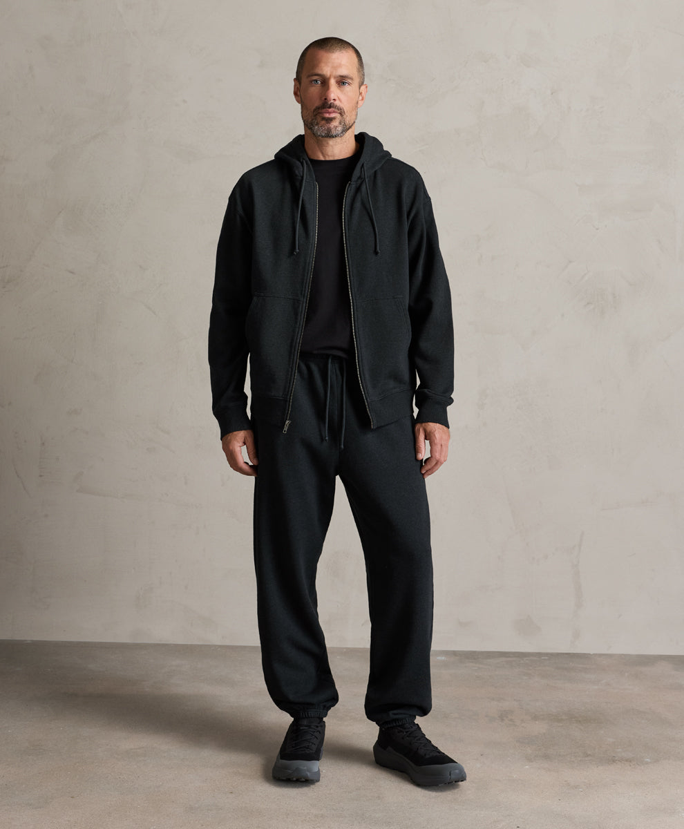 A man wearing a Pact charcoal heather zip-up hoodie, sweatpants, and black sneakers stands against a textured light gray concrete wall.