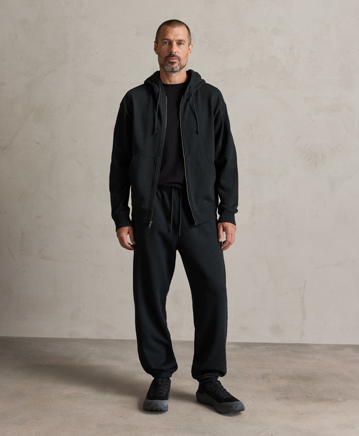A man wearing a Pact charcoal heather zip-up hoodie, sweatpants, and black sneakers stands against a textured light gray concrete wall.