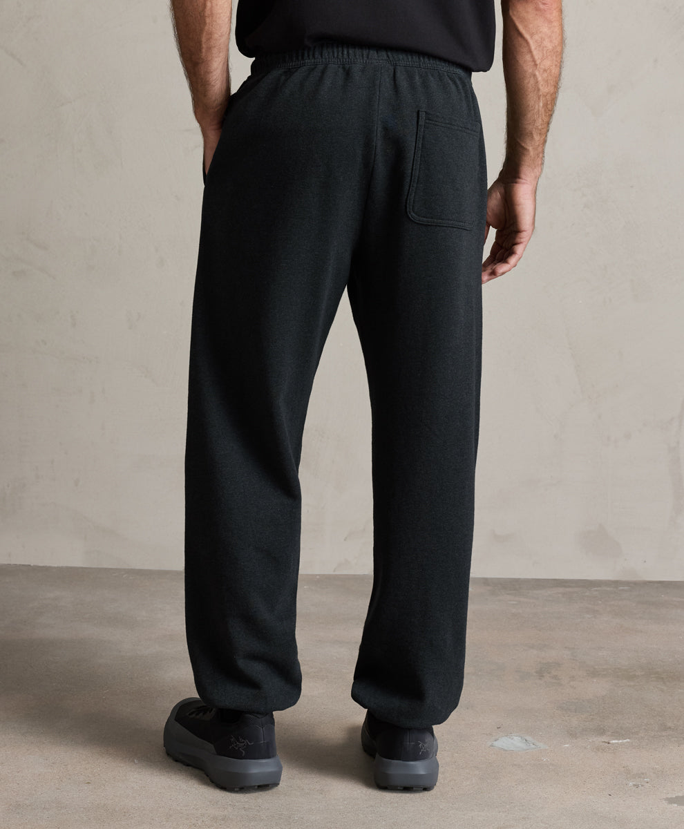 A person shown from the back wearing loose charcoal heather drawstring sweatpants with a side pocket, paired with a black top and black shoes, standing against a textured beige wall and concrete floor.