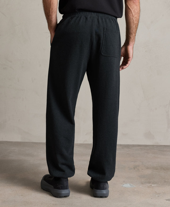 A person shown from the back wearing loose charcoal heather drawstring sweatpants with a side pocket, paired with a black top and black shoes, standing against a textured beige wall and concrete floor.