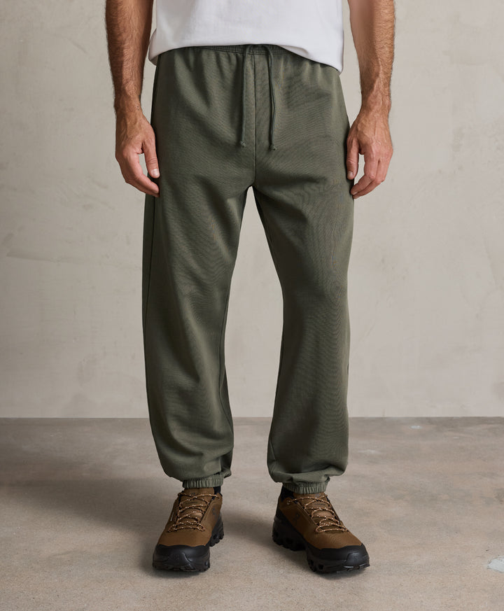 A person wearing pact dusted green drawstring jogger-style pants with elastic cuffs, paired with brown hiking boots and a white top, standing against a textured beige wall and concrete floor.