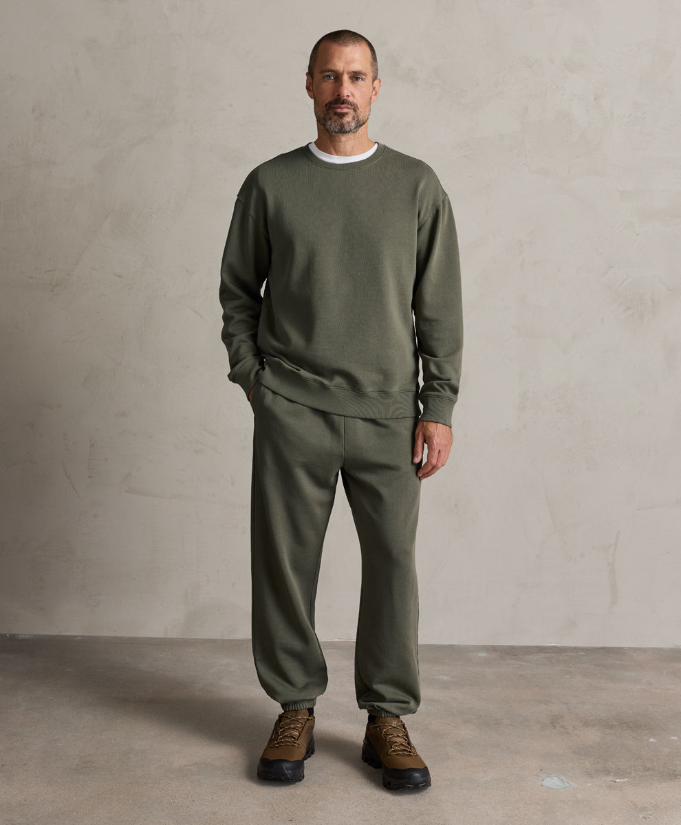 A man wearing a dusted green crewneck sweatshirt and matching sweatpants, paired with brown lace-up shoes, standing against a textured light gray studio wall.
