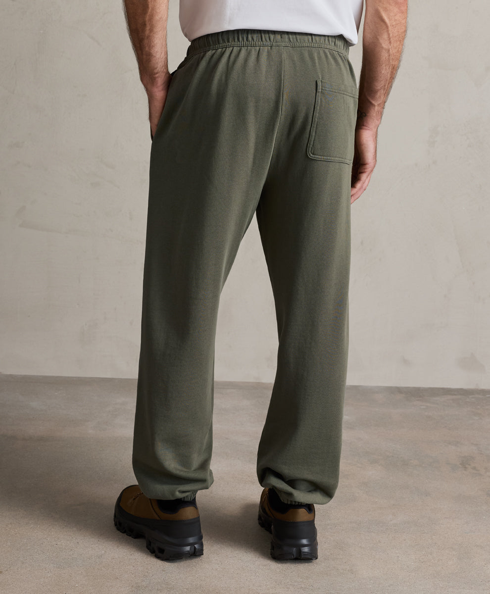 A person shown from the back wearing loose dusted green sweatpants, a white top, and brown shoes, standing against a textured beige wall and concrete floor.