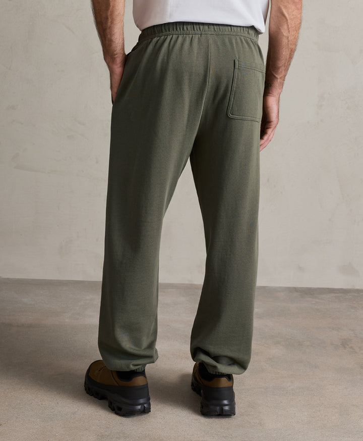A person shown from the back wearing loose dusted green sweatpants, a white top, and brown shoes, standing against a textured beige wall and concrete floor.