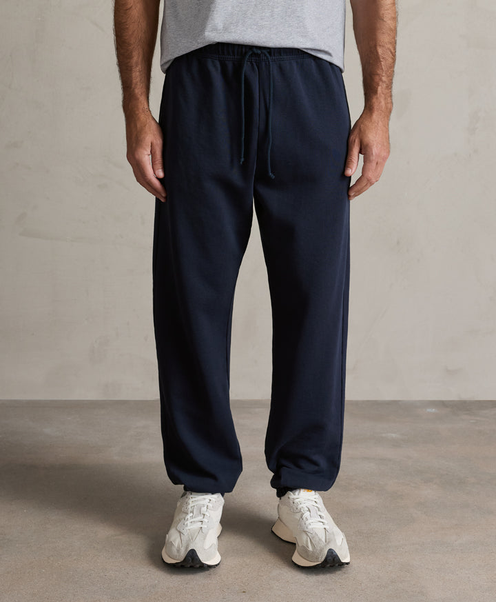 A person wearing pact dark navy drawstring sweatpants, a light gray t-shirt, and white sneakers, standing against a neutral concrete-textured wall.