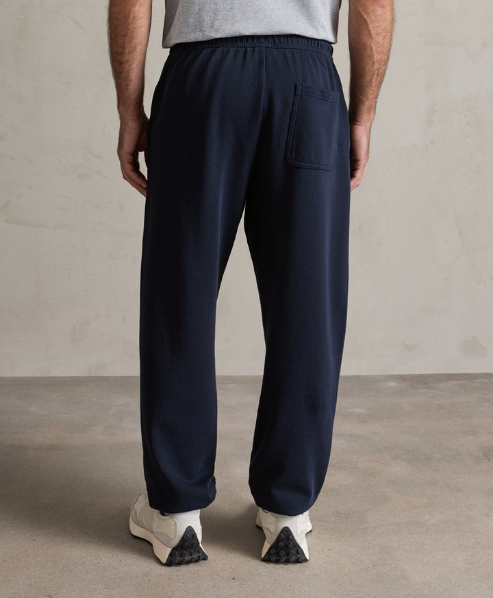 A person shown from the back wearing dark navy jogger-style pants with an elastic waistband, side pocket, and relaxed straight-leg fit, paired with a light gray top and white sneakers against a neutral indoor backdrop.