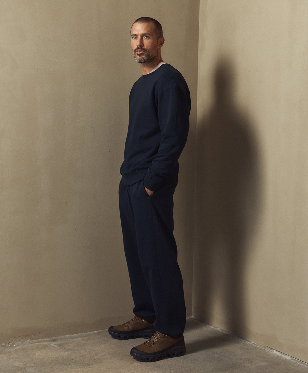 A man stands in profile wearing a dark navy crewneck sweatshirt and matching navy jogger pants, paired with brown hiking shoes, against a textured beige plaster wall corner.