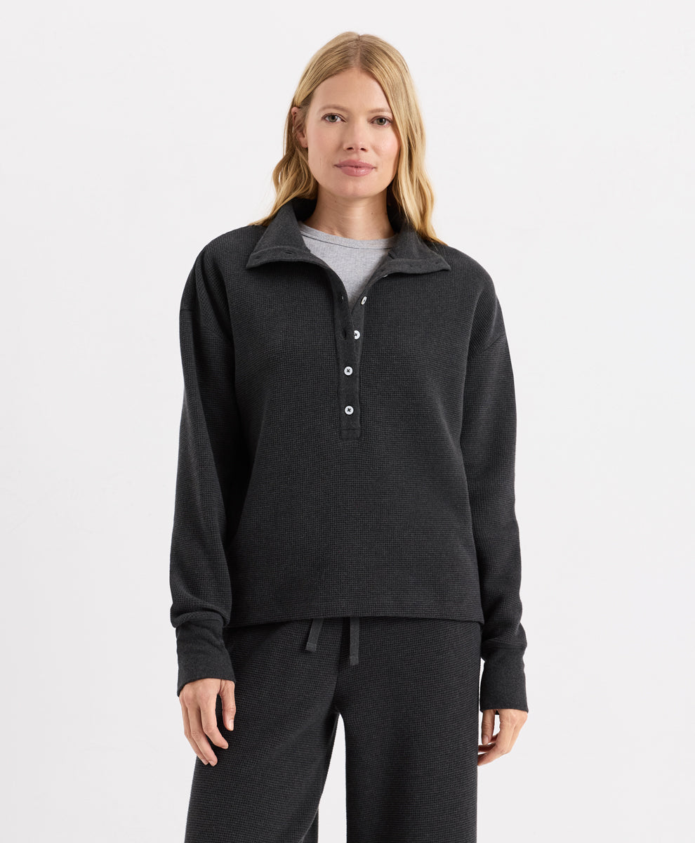 A woman modeling a Pact dark charcoal heather long-sleeve quarter-placket henley top with white buttons, paired with matching dark sweatpants and a light gray undershirt, against a plain white studio background.