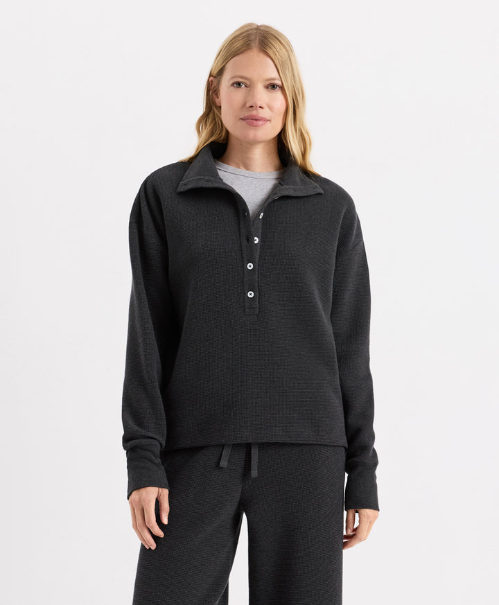 A woman modeling a Pact dark charcoal heather long-sleeve quarter-placket henley top with white buttons, paired with matching dark sweatpants and a light gray undershirt, against a plain white studio background.