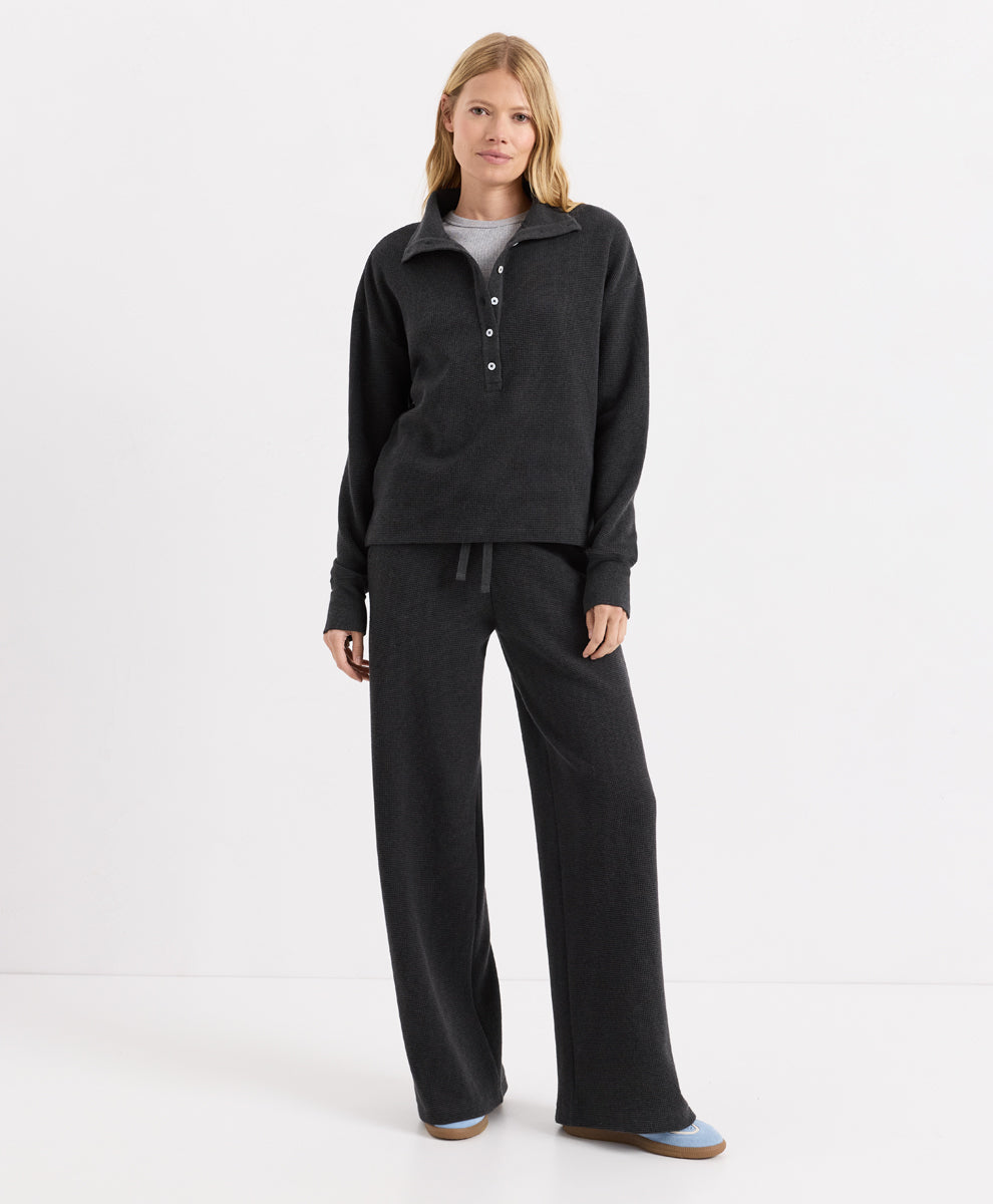 A woman modeling a dark charcoal heather two-piece loungewear set consisting of a long-sleeve buttoned henley pullover and matching wide-leg pants, photographed against a seamless white studio background.