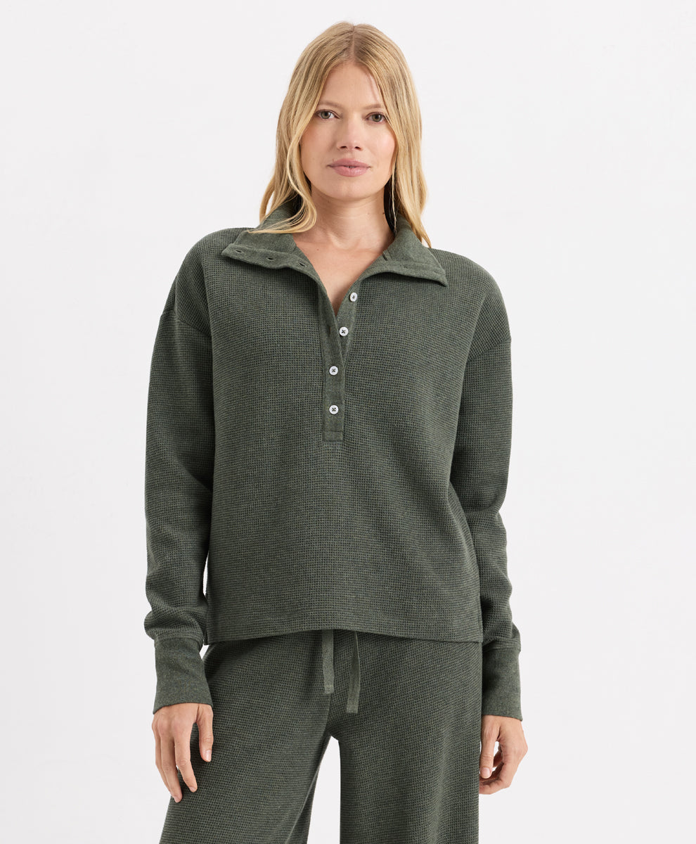 A woman modeling a dusted green heather knit loungewear set, featuring a long-sleeve henley-style top with matching drawstring sweatpants, against a plain light gray studio background.