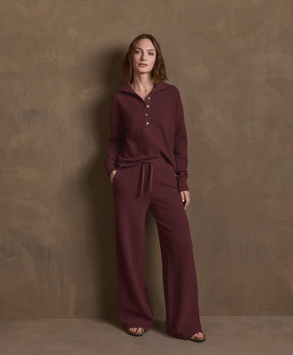 A woman stands against a textured brown studio backdrop wearing a matching merlot heather knit loungewear set consisting of a long-sleeve henley top with a drawstring and wide-leg pants.