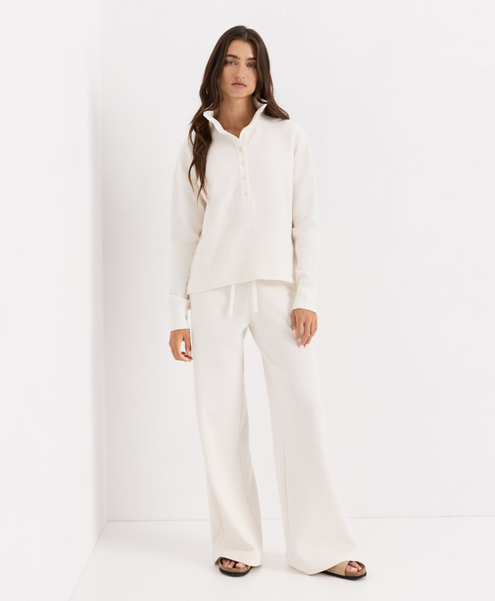 A woman modeling a matching soft ivory lounge set consisting of a long-sleeve henley and wide-leg trousers, standing against a clean white studio background.