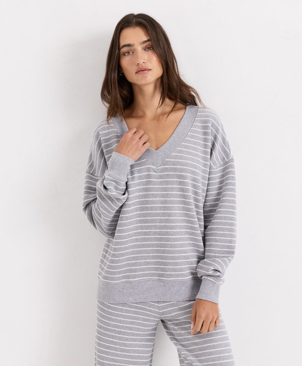 A woman modeling a light gray and white striped long-sleeve V-neck top with matching striped pants against a plain white studio background.