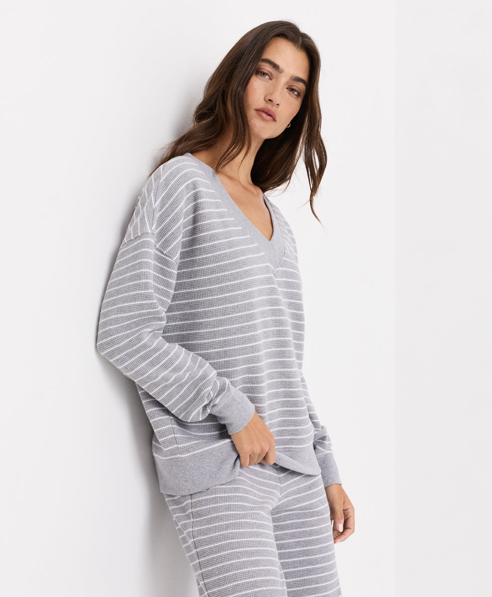 A woman wearing a light gray and white striped long-sleeve V-neck top and matching striped pants, posing against a plain white wall.
