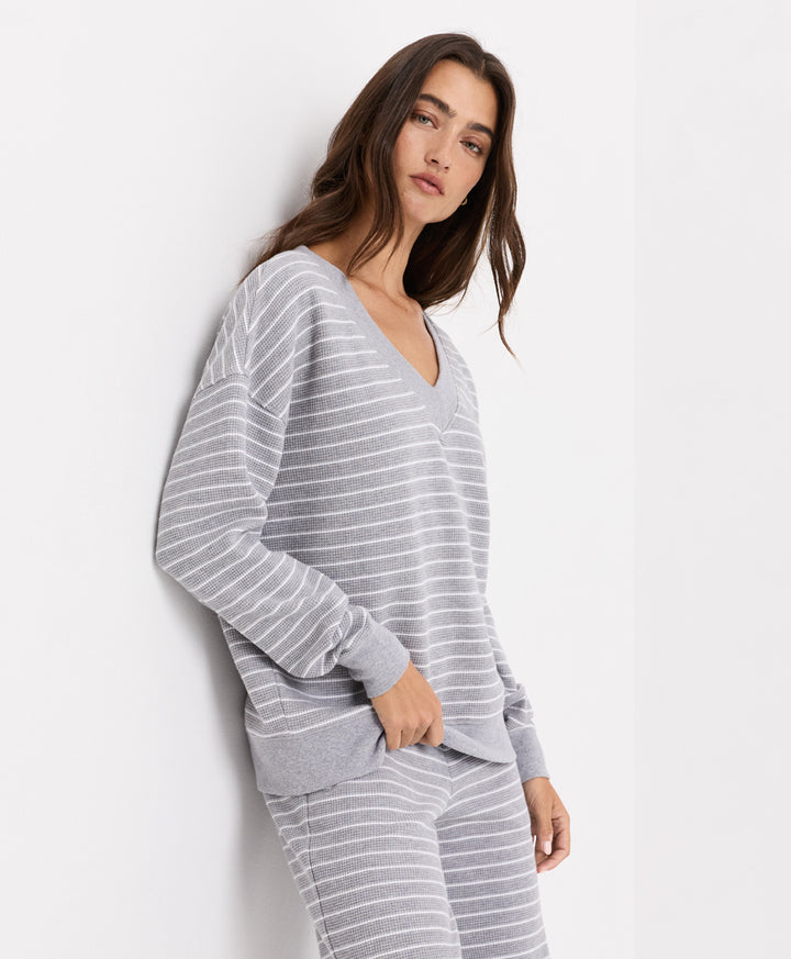 A woman wearing a light gray and white striped long-sleeve V-neck top and matching striped pants, posing against a plain white wall.