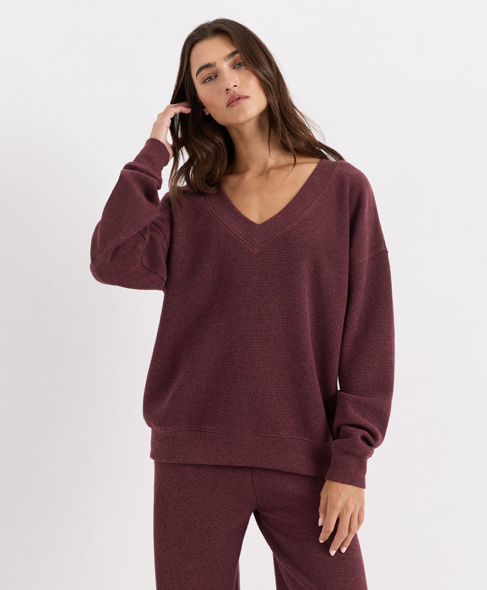 A woman modeling a matching merlot heather lounge set consisting of a V-neck long-sleeve top and coordinating pants, shot against a plain light gray studio background.