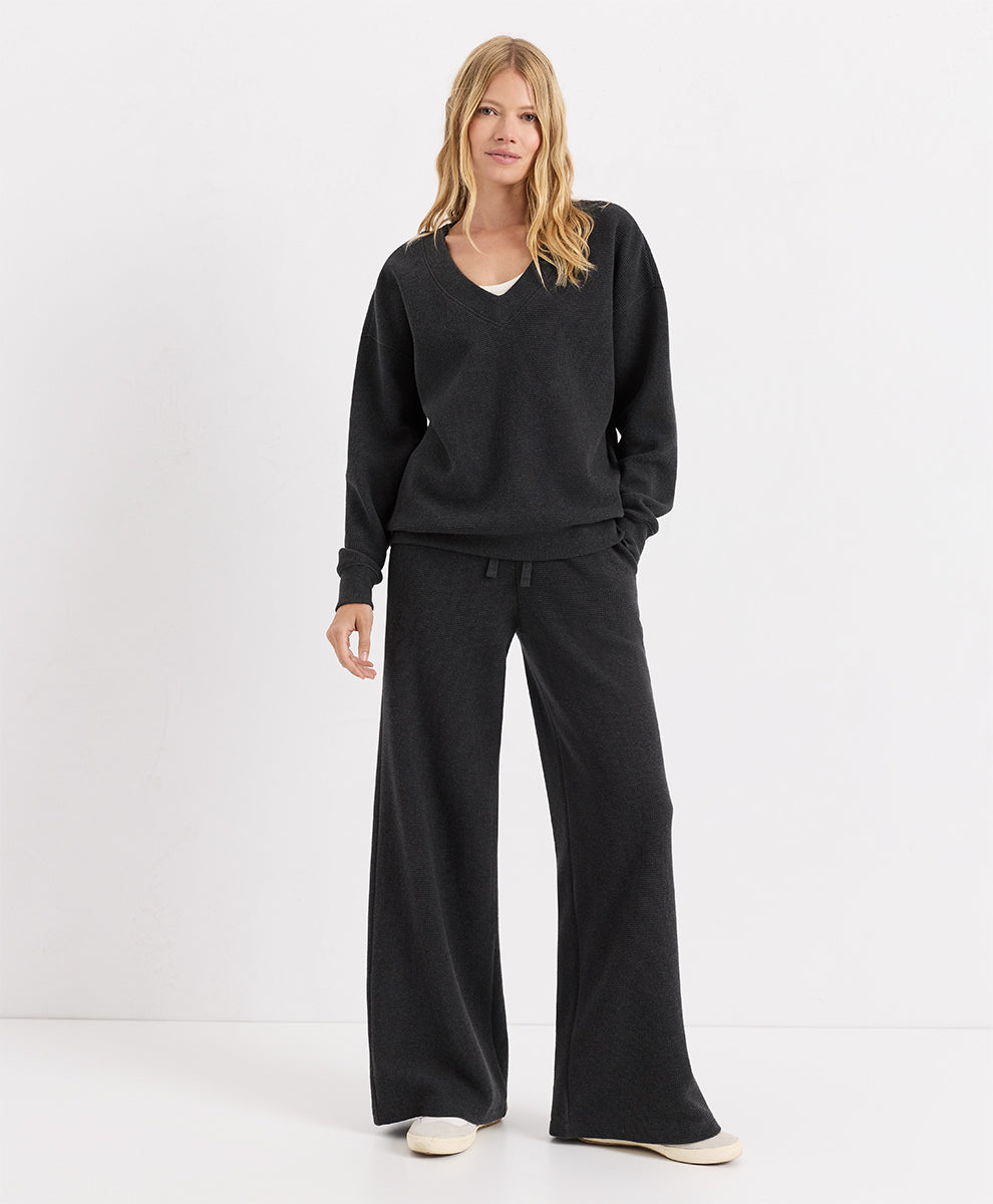 A woman modeling a dark charcoal heather lounge set consisting of a V-neck long-sleeve top and matching wide-leg pants, paired with off-white slip-on shoes, against a plain white studio background.