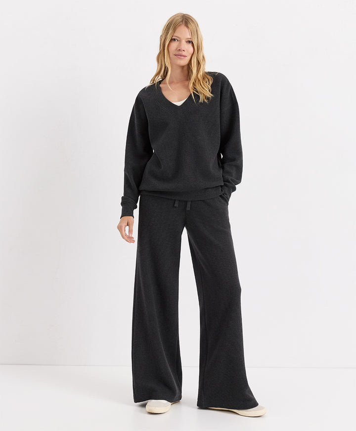 A woman modeling a dark charcoal heather lounge set consisting of a V-neck long-sleeve top and matching wide-leg pants, paired with off-white slip-on shoes, against a plain white studio background.