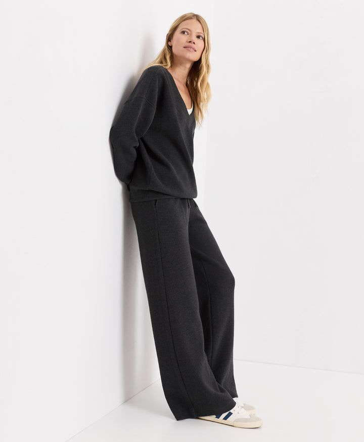A woman wearing a dark charcoal heather knit lounge set consisting of a V-neck top and matching wide-leg pants, leaning against a white wall in a minimalist studio setting.